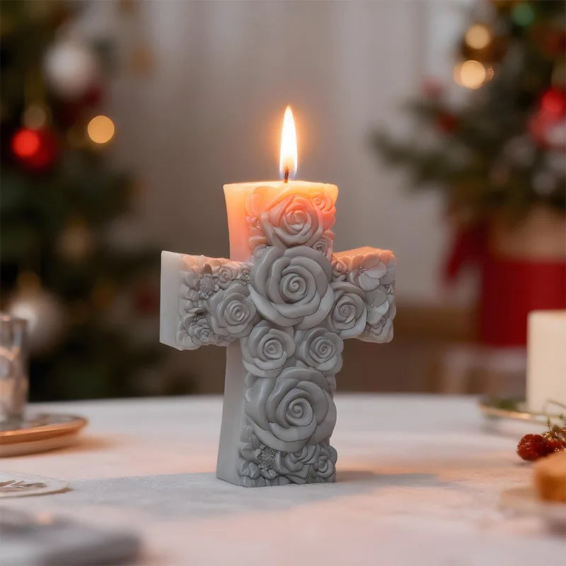 A white floral cross candle with a lit wick sits on a table in a softly lit holiday setting.