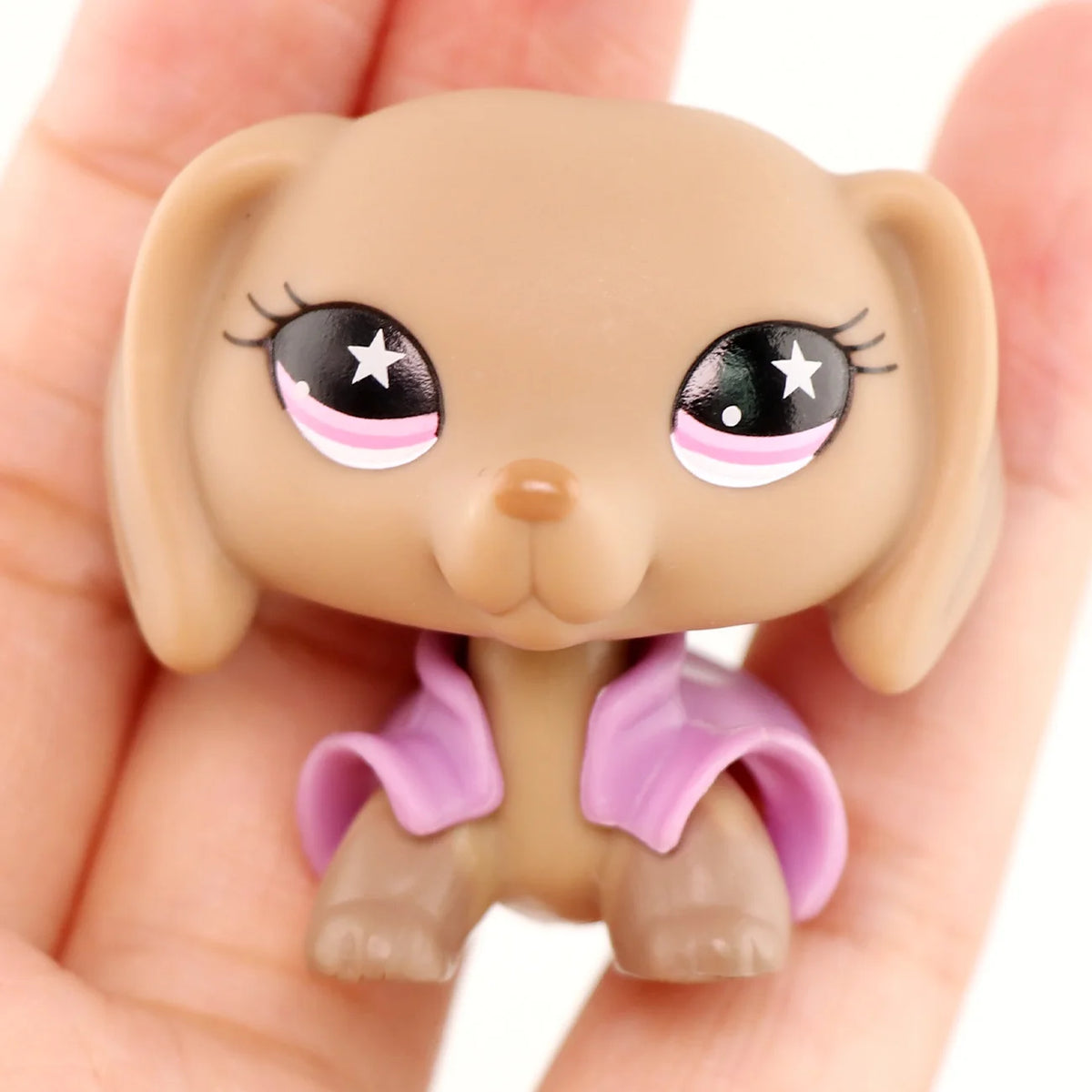 A small, light brown miniature pet animal figure with large eyes and a purple accessory is held in a hand.