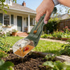 A hand holds a green handheld insect catcher with a magnifying lens over soil in a garden.