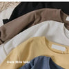 A stack of three kids' solid color long sleeve cotton tees in beige, white, and yellow.