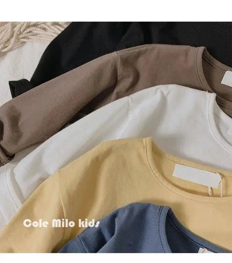 A stack of three kids' solid color long sleeve cotton tees in beige, white, and yellow.