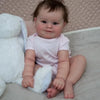 A baby reborn doll named Maddie is sitting on a light surface, wearing a pink shirt and holding a white cloth.