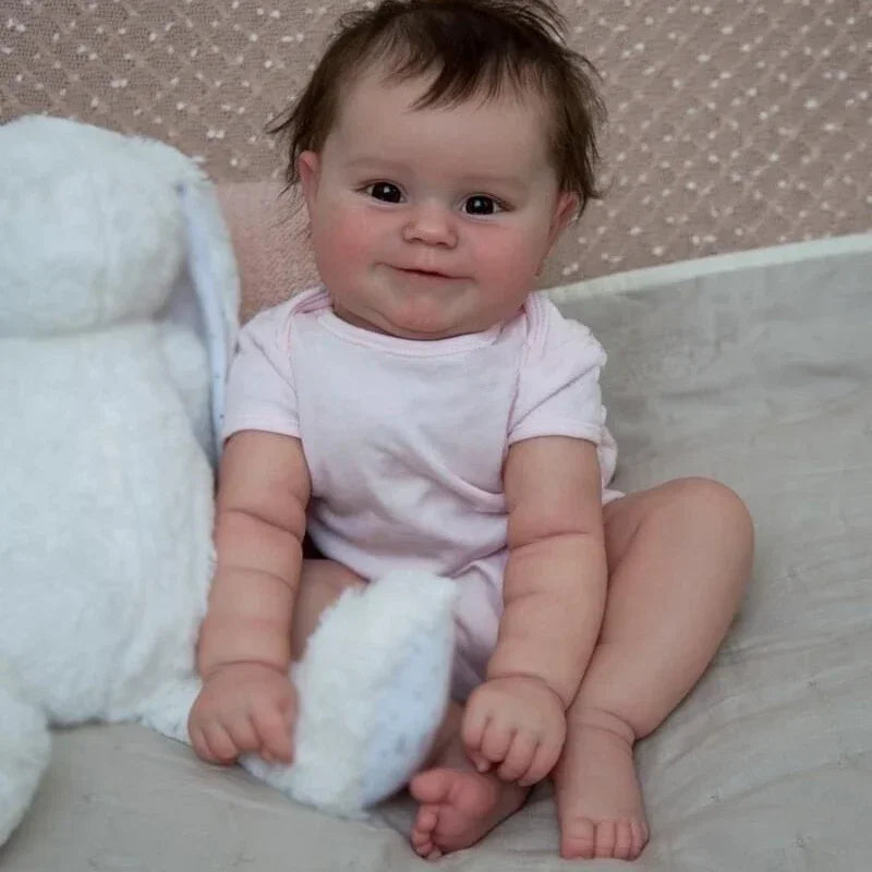 A baby reborn doll named Maddie is sitting on a light surface, wearing a pink shirt and holding a white cloth.