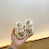 A hand holds a pair of white soft sole sandals for baby's first steps featuring a cute bear design.