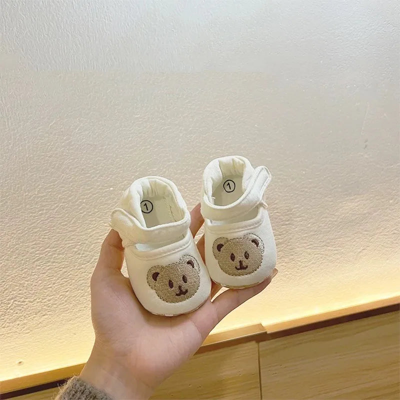 A hand holds a pair of white soft sole sandals for baby's first steps featuring a cute bear design.