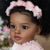 A close-up portrait of a toddler doll with hand-rooted black hair adorned with pink floral headpiece.