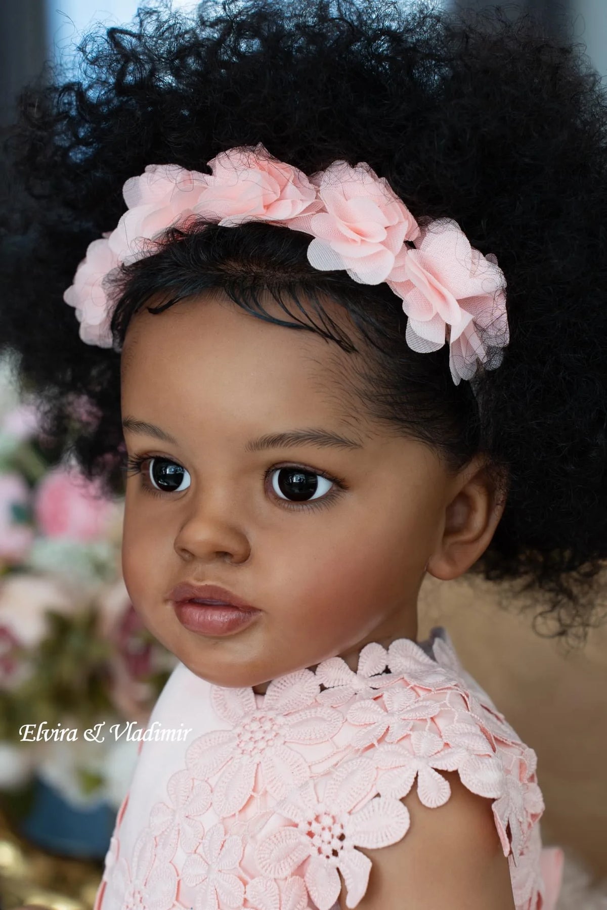A close-up portrait of a toddler doll with hand-rooted black hair adorned with pink floral headpiece.