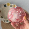 A hand holds a pink, translucent stress ball filled with glitter and small stars against a light background.