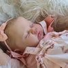 A newborn baby doll with hand-painted skin is sleeping peacefully on a white, fluffy surface.