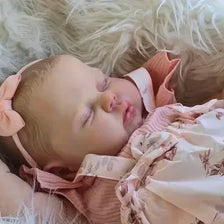 A newborn baby doll with hand-painted skin is sleeping peacefully on a white, fluffy surface.