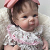 A lifelike 18-inch reborn baby doll with hand-painted details wearing a pink floral dress and a pink bow, resting on a white 
