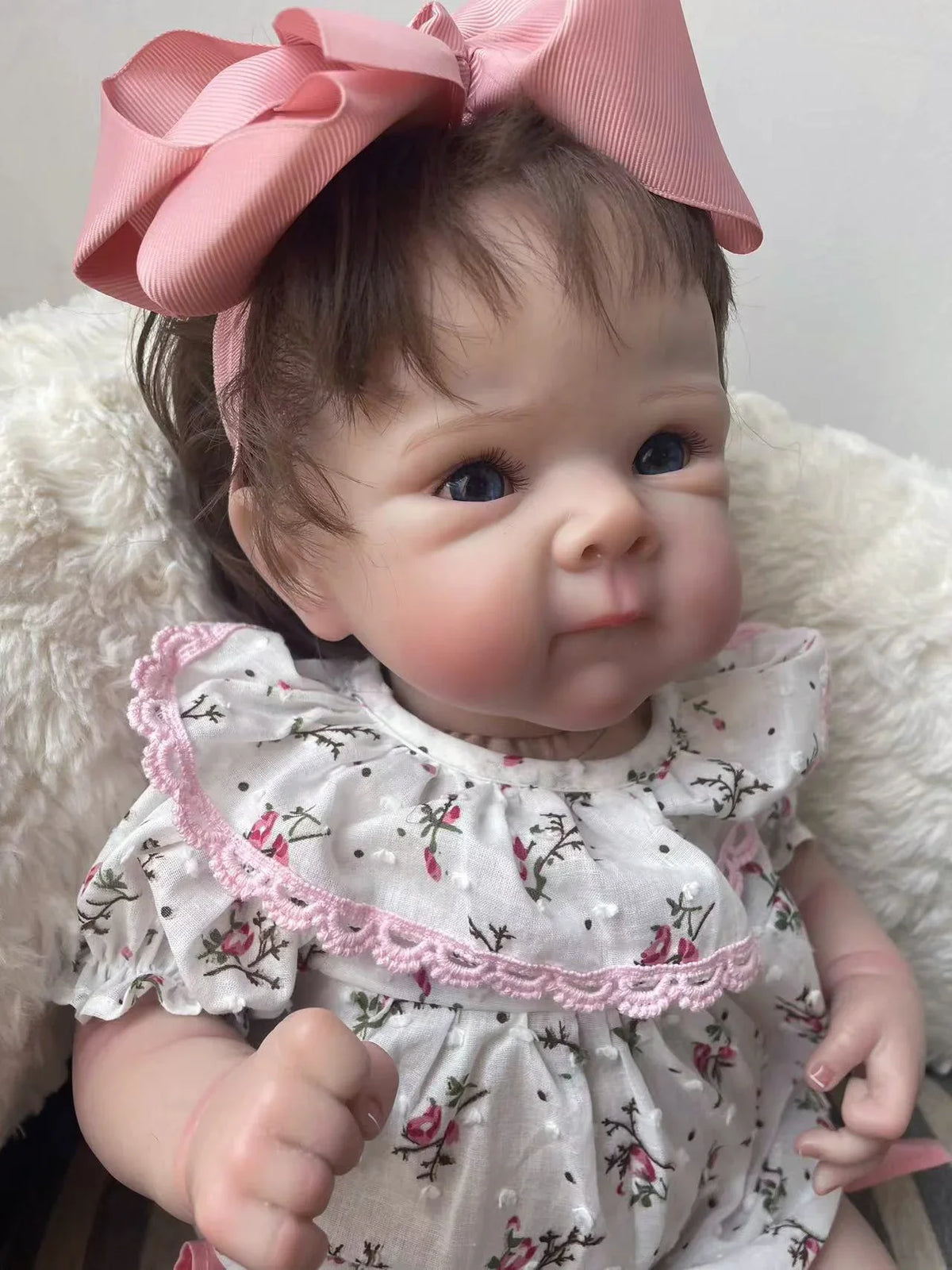 A lifelike 18-inch reborn baby doll with hand-painted details wearing a pink floral dress and a pink bow, resting on a white 