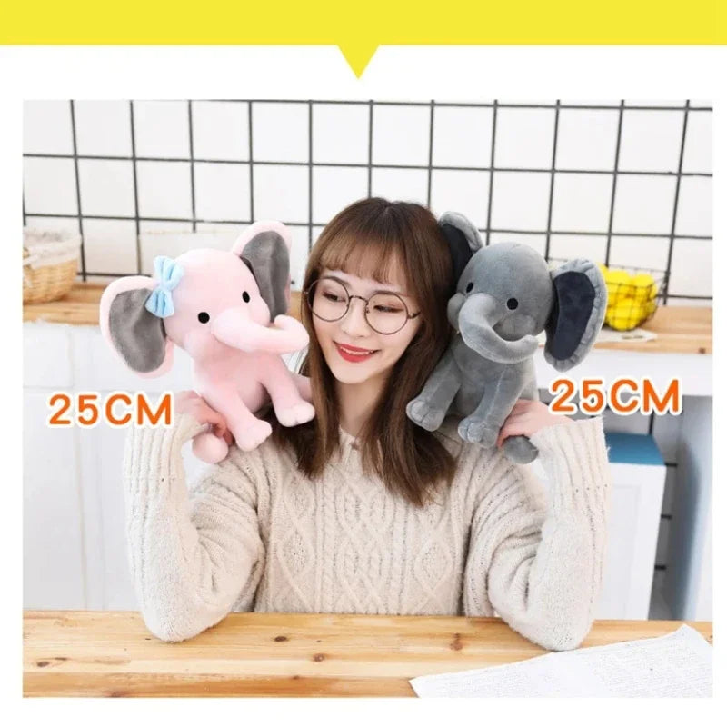 A woman is holding a gray elephant plush toy, demonstrating its size against a measuring tape.