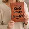 A person holds a brown leather-bound Bible study guide with gold lettering and an embossed design.
