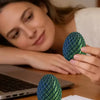 A woman is holding a teal dragon egg fidget spinner next to a laptop and some papers.
