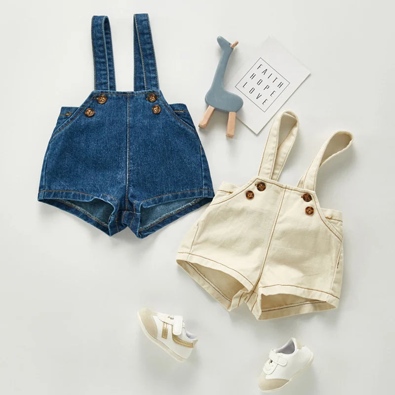 A pair of denim overalls or romper with pockets and wooden buttons, displayed on a light surface.