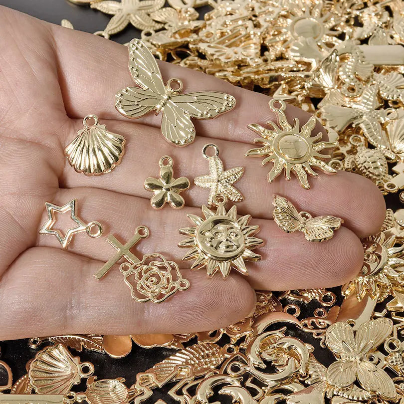 A hand holds various gold-toned mixed metal charms including a shell, flower, star, and sunburst designs.