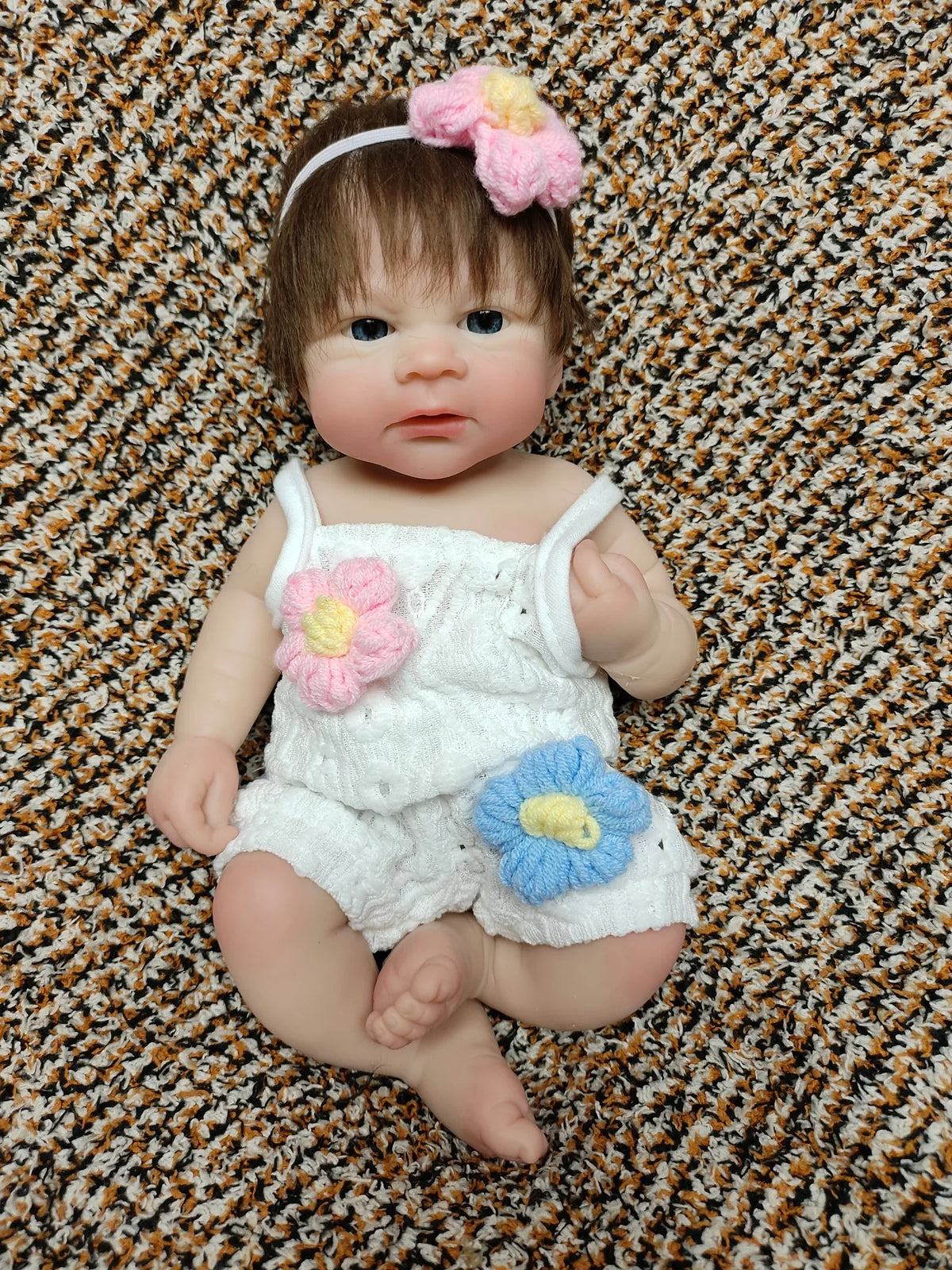 A realistic soft silicone reborn doll with brown hair and pink floral accessories sitting on a textured brown surface.
