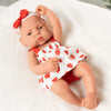 A lifelike vinyl reborn doll with a red bow and a strawberry print outfit lying on a white surface.