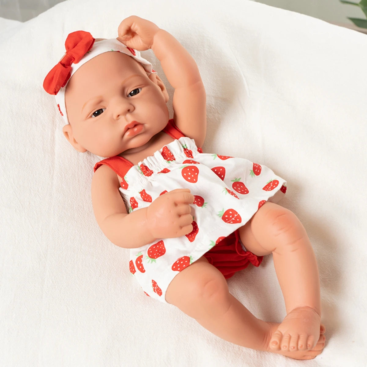 A lifelike vinyl reborn doll with a red bow and a strawberry print outfit lying on a white surface.