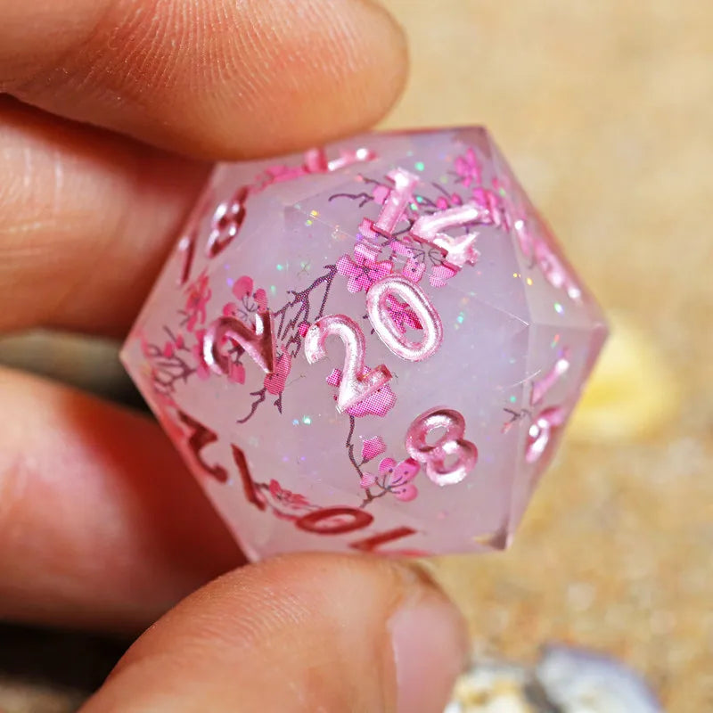 A hand holds a pink hexagonal resin polyhedral die featuring pink floral designs and the number 2023.