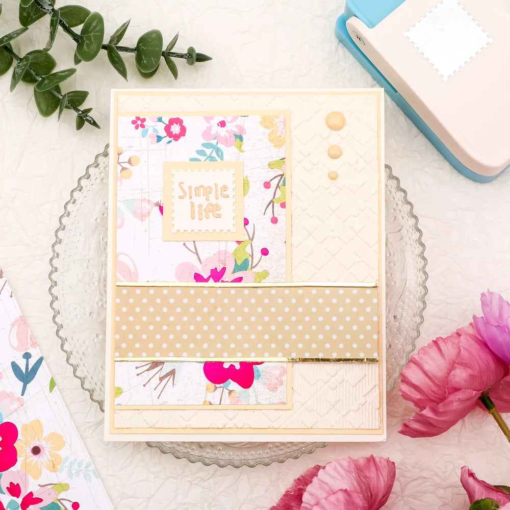 Un perforateur de timbres carré avec un design rose pâle et crème, posé sur une surface blanche.