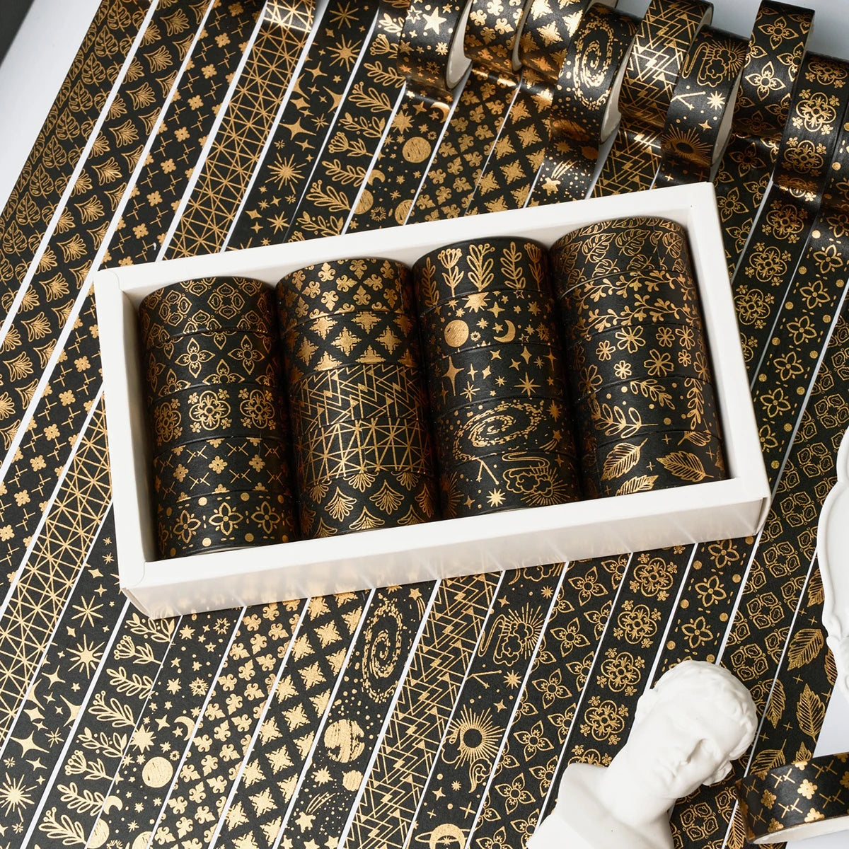 A set of 20 rolls of black and gold foil washi tape displayed in a white box on a patterned surface.