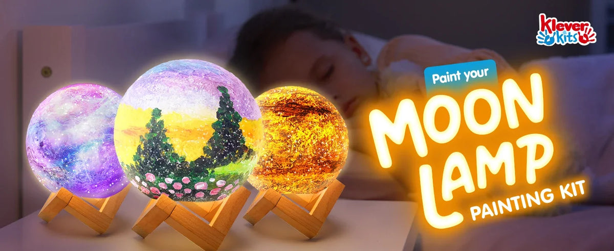 A close-up of a DIY Moon Lamp painting kit featuring three glowing spherical night lights resting on a wooden surface.