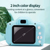 A light blue mini digital camera with a black screen is displayed on a white surface next to a green plant.
