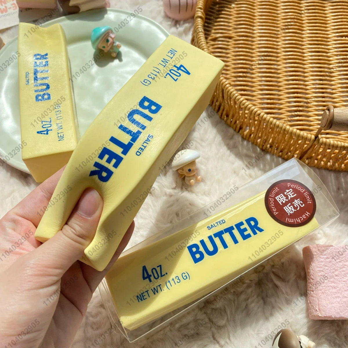 A hand holds a yellow butter stick squishy toy with a blue label and a small wooden figurine in the background.
