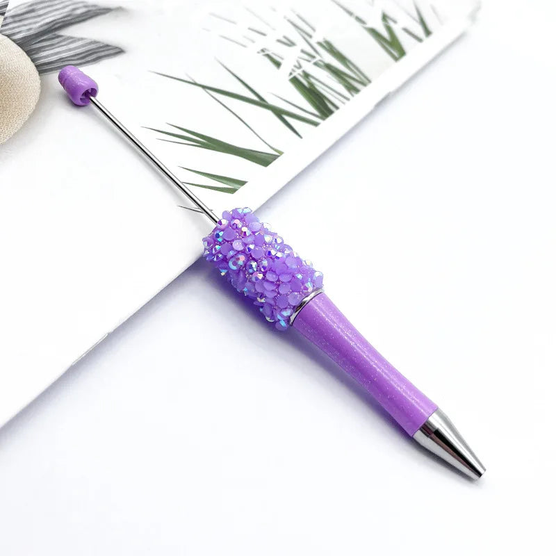 A purple ballpoint pen with a glittery purple bead section rests on a white background next to green foliage.