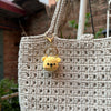 A light beige crocheted bag features a small yellow teddy bear keychain attached to the strap.