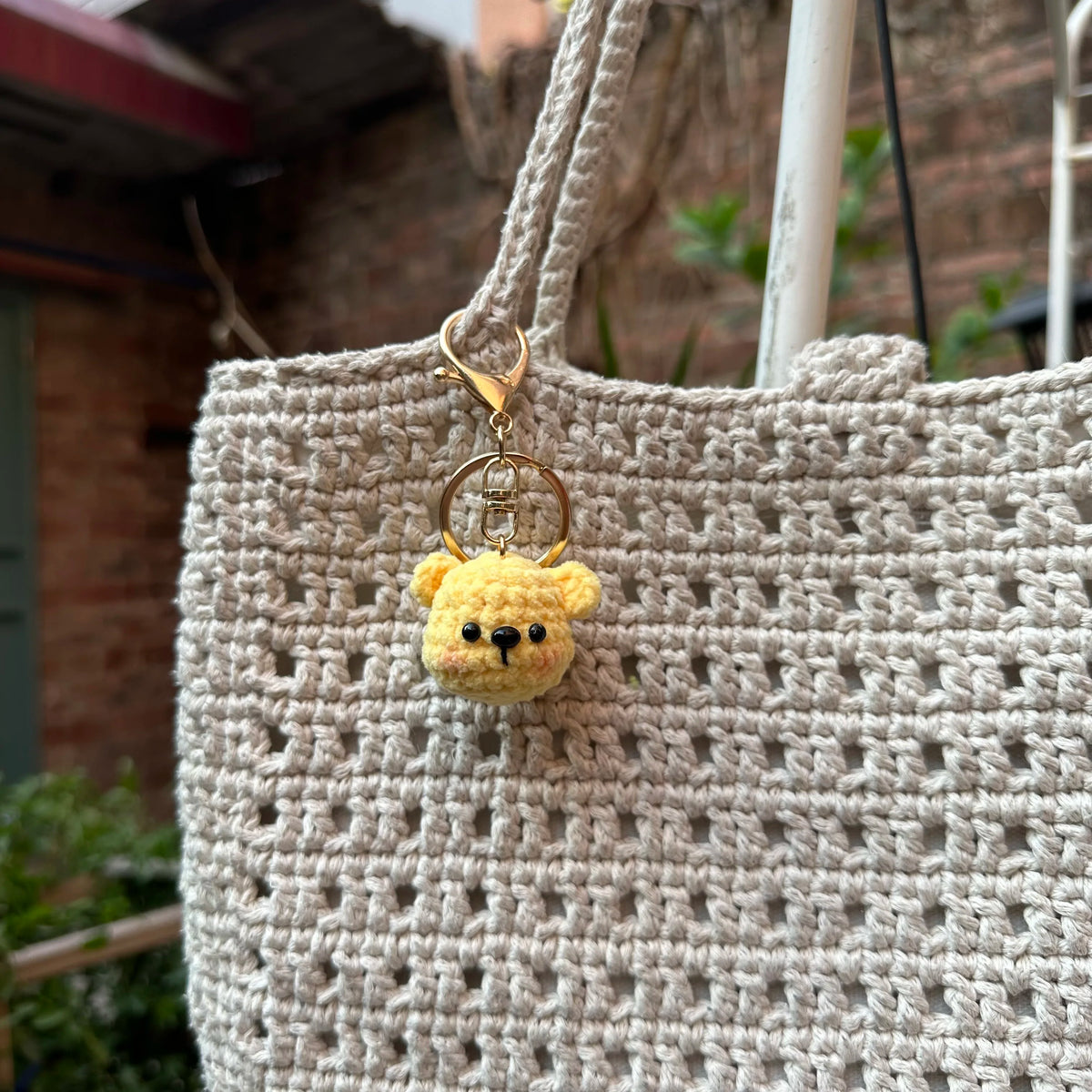 A light beige crocheted bag features a small yellow teddy bear keychain attached to the strap.