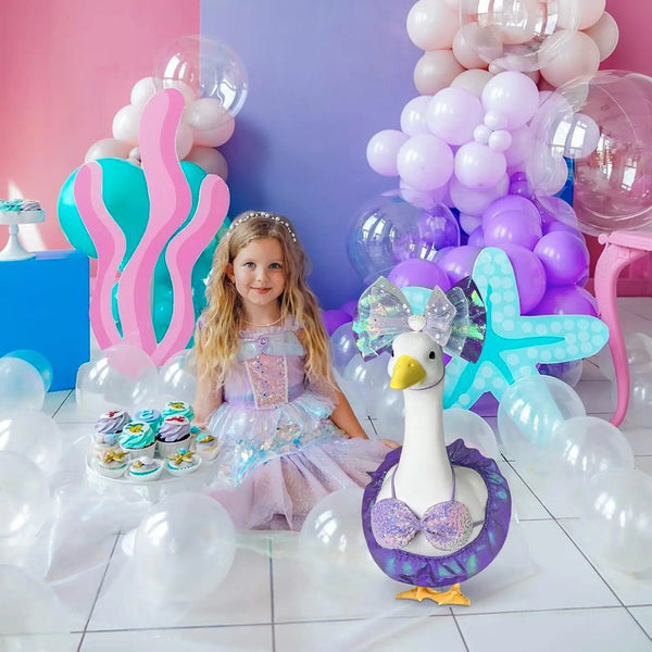 Ein junges Mädchen in einem lila Meerjungfrauen-Ganskostüm steht vor pinken und lila Ballons.