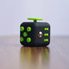 A black fidget cube with green buttons and a silver sphere resting on a wooden surface.