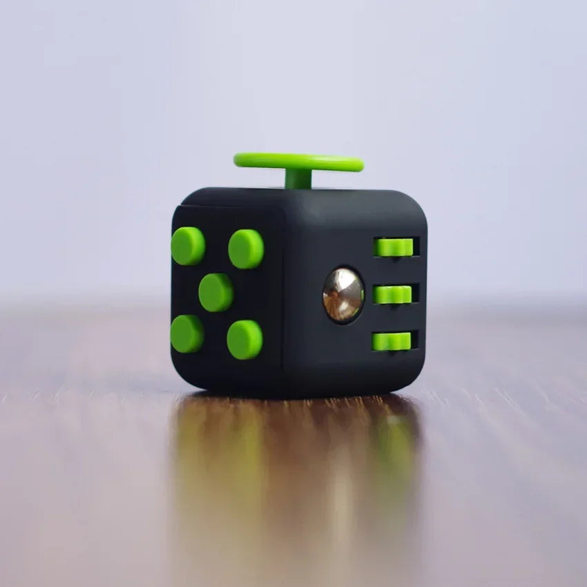 A black fidget cube with green buttons and a silver sphere resting on a wooden surface.