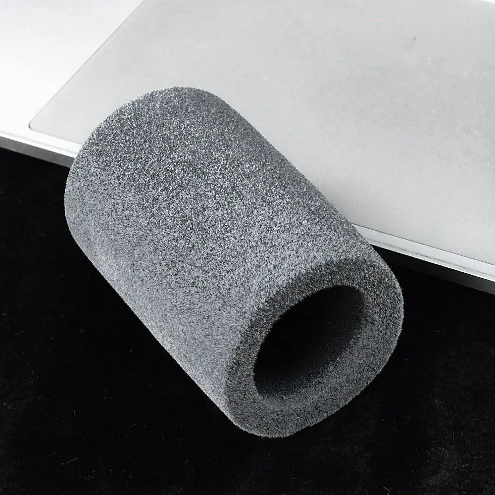 A cylindrical flocked foam microphone windscreen cover resting on a dark surface next to a white object.