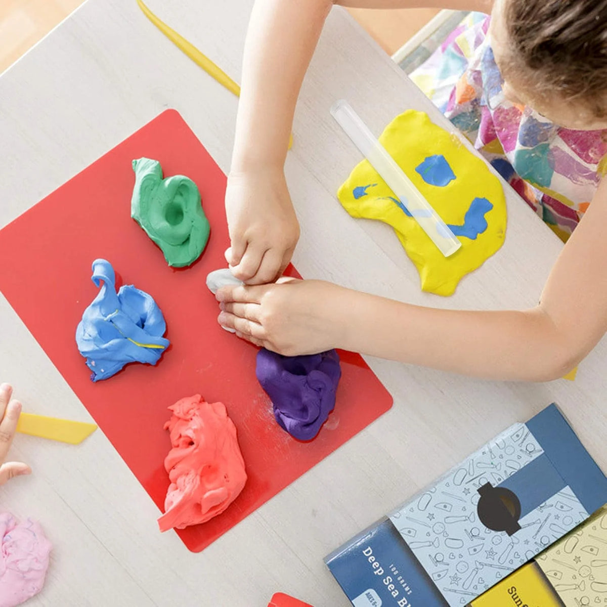 A child is using an acrylic clay roller and sheet set to sculpt various colored clay pieces on a red surface.
