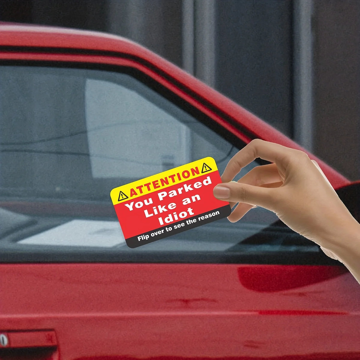 A hand holds up a funny parking violation card with text warning you parked like an idiot against a red car.