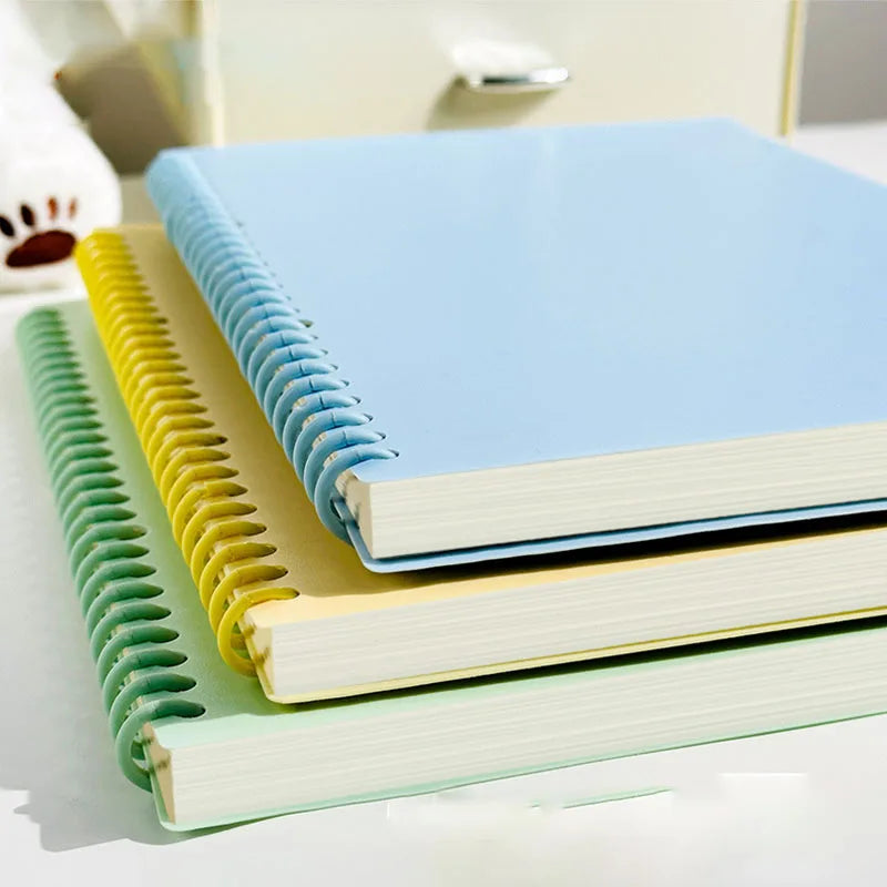 A stack of pastel blue and green spiral notebooks with yellow covers resting on a white surface.
