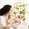 A woman gently interacts with a baby in a crib surrounded by a wooden felt forest animal mobile.