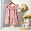 A pink floral smocked summer dress for girls with flying sleeves displayed on a wooden hanger.