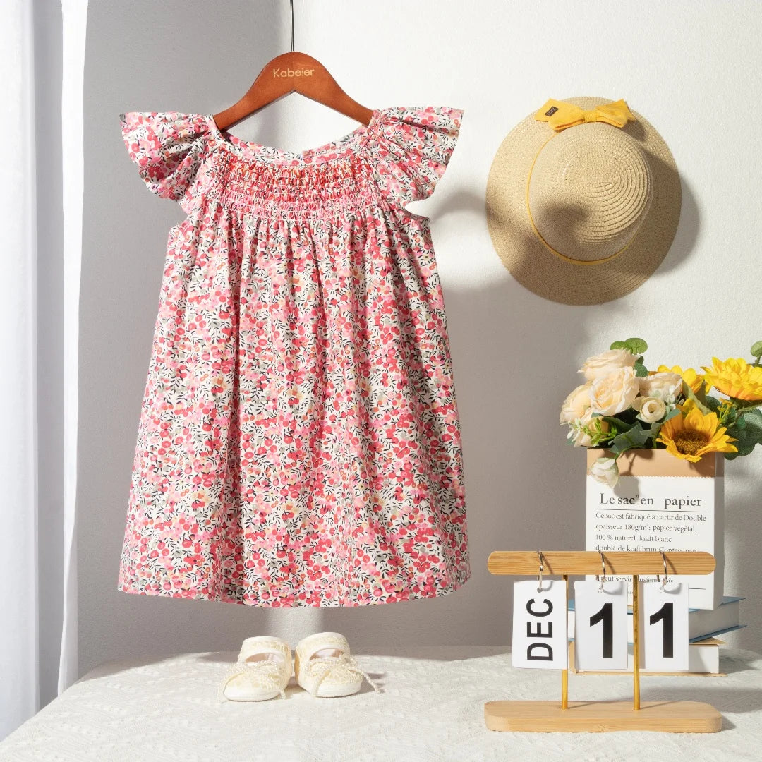 A pink floral smocked summer dress for girls with flying sleeves displayed on a wooden hanger.
