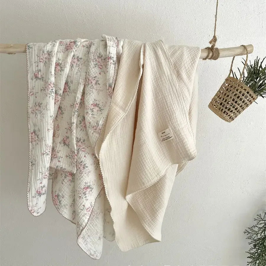 A cream colored muslin swaddle blanket with a pink floral print is hanging on a wooden rod.