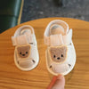 A pair of light pink soft sole first walker shoes shaped like bears resting on a wooden surface.