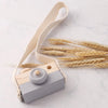 A wooden camera teether toy with a light wood body and gray button, attached to a beige fabric strap, resting on a white marb