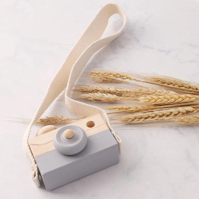 A wooden camera teether toy with a light wood body and gray button, attached to a beige fabric strap, resting on a white marb