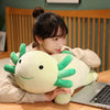 A person is holding a large, light green axolotl plush pillow while sitting at a wooden table.