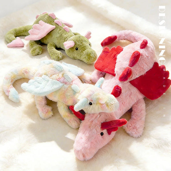 A collection of three soft plush dragon toys in green, yellow, and pink resting on a white fluffy surface.