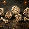 A set of bone and skull carved polyhedral dice resting on a dark wooden surface surrounded by lit candles.
